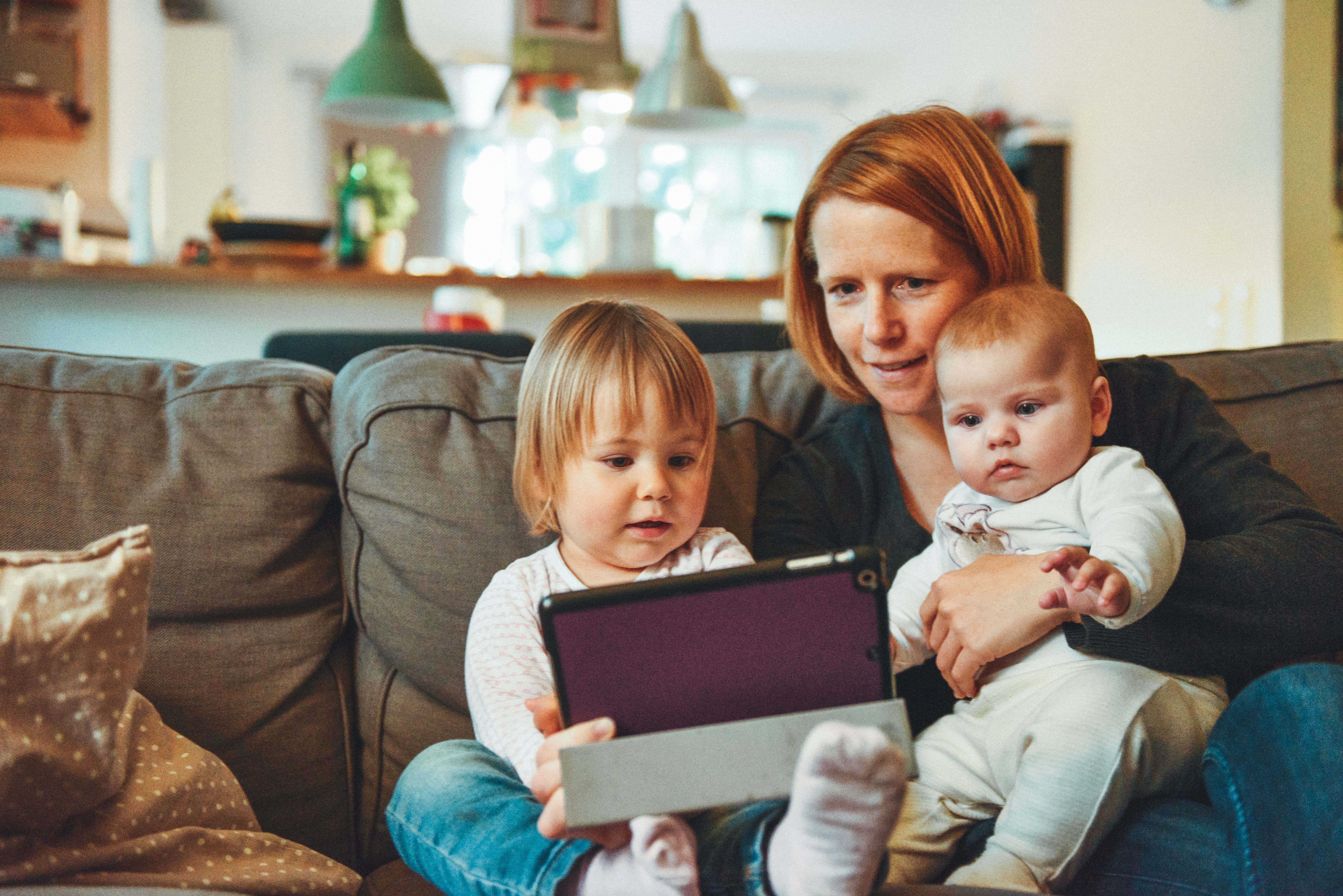 Blonde woman holding baby next to a toddler playing with a tablet.
